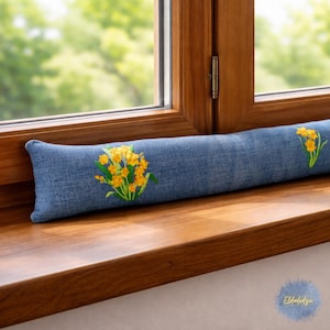 Può includere: Un paraspifferi in denim blu con fiori gialli ricamati. Il lungo cuscino rettangolare è posto su un davanzale di legno. I fiori sono raggruppati in due punti.