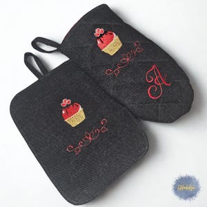 Può includere: Set di presina e guanto da forno in denim nero. Ogni pezzo presenta un cupcake ricamato in rosso con dettagli dorati e una scritta decorativa rossa. Il guanto da forno ha la lettera "A" ricamata in rosso. Anello nero per appendere.
