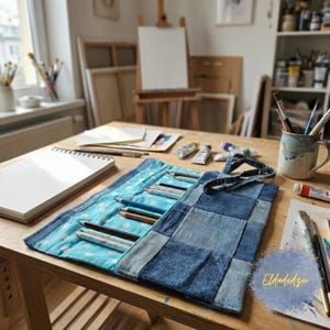 Puede incluir: Un estuche enrollable para lápices de mezclilla y lunares azules con asa, que contiene varios lápices de colores. El rollo está abierto, mostrando los lápices, y está sobre una mesa de madera con materiales de arte y un cuaderno de bocetos.