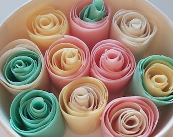 May include: A white bowl filled with colorful rolled ice cream. The ice cream is rolled into a rose shape and comes in a variety of colors, including pink, yellow, green, and white.