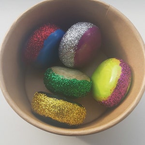 Peut inclure: Cinq pierres rondes peintes de différentes couleurs et recouvertes de paillettes. Les pierres sont dans un bol en papier brun. Les couleurs sont le rouge, l'argent, le violet, le vert et l'or.