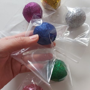 May include: Six small, round, glitter-covered ornaments in individual plastic bags. The ornaments are pink, blue, green, gold, silver, and a lighter shade of pink.