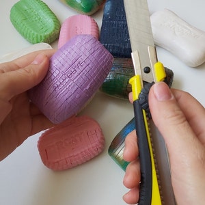May include: A selection of colourful patterned soaps are arranged on a white surface. A yellow and black utility knife is being used to cut a purple soap bar.