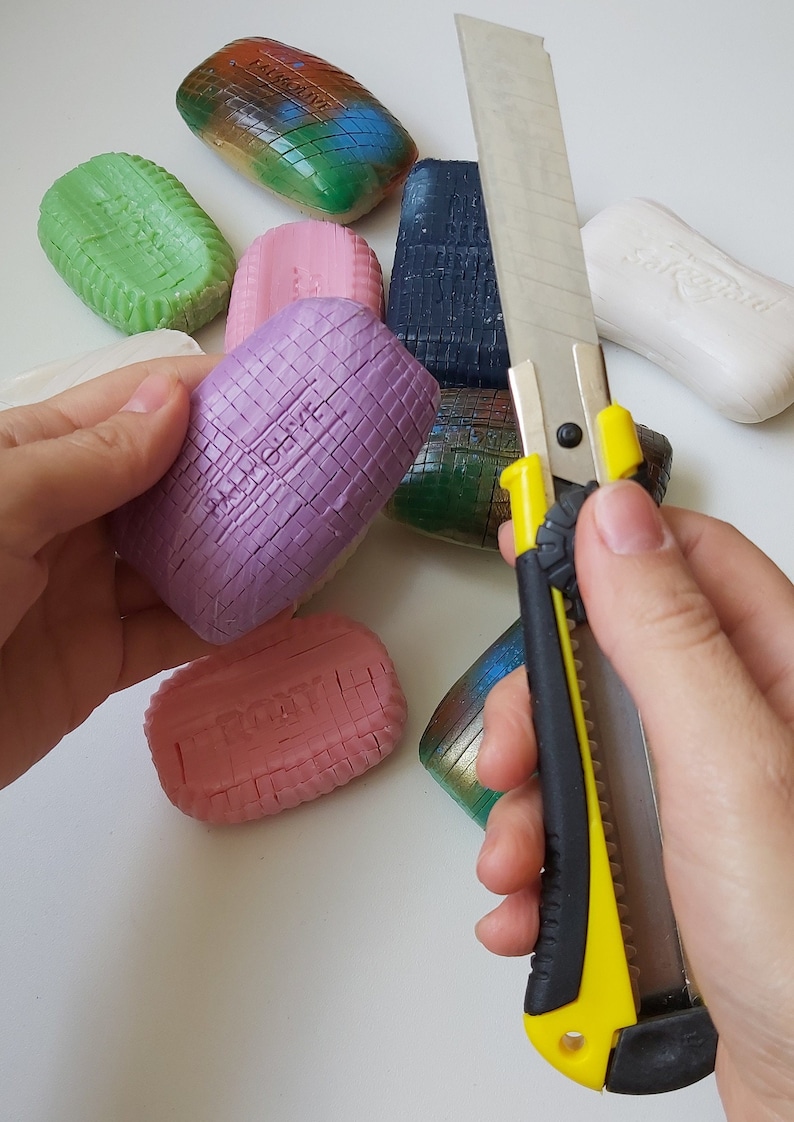 May include: A selection of colourful patterned soaps are arranged on a white surface. A yellow and black utility knife is being used to cut a purple soap bar.