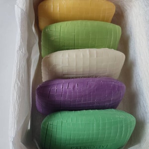May include: Five bars of soap stacked on top of each other. The bars are all different colours: yellow, green, white, purple, and green. The bars are all rectangular and have a grid pattern on them. The bottom bar has the word "PALMOLIVE" printed on it.
