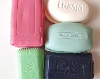 May include: An assortment of six soaps in various shapes and colors. The soaps include a pink rectangular bar, a white oval bar with the word "Luksja", a light green rectangular bar with the word "Shik", a blue-green rectangular bar with the word "Palmolive", and a dark blue rectangular bar.
