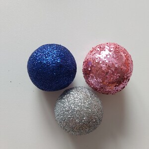 May include: Three round Christmas ornaments with glitter. One is blue, one is pink, and one is silver.