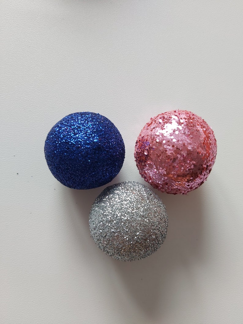May include: Three round Christmas ornaments with glitter. One is blue, one is pink, and one is silver.