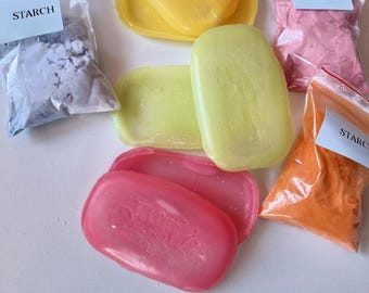 May include: An assortment of colorful soaps and bags of starch. The soaps are yellow, green, and pink, with a floral design on the pink ones. The bags contain blue, pink, and orange powders, each labeled "STARCH".