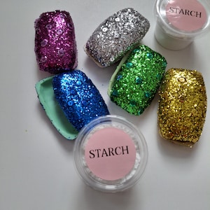 May include: Six small containers filled with glitter in various colors, including pink, silver, green, blue, and gold. Two containers of white powder labeled "STARCH" are also included.
