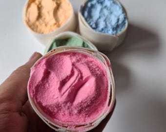 May include: Four small containers of colorful sand, one pink, one blue, one green, and one orange. The pink container is being held in a hand.