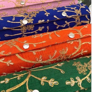 May include: A close-up of several fabric boxes in pink, blue, orange, and green. Each box features gold floral embroidery and a silver button closure. The boxes are likely for storage or decorative purposes. The boxes are rectangular in shape.