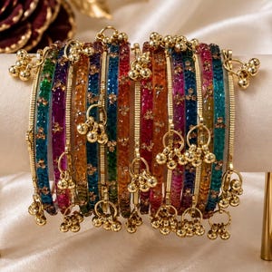 May include: A set of multi-coloured translucent bangles featuring a textured surface with embedded gold-tone accents. Each bangle is adorned with small, dangling gold-tone metal bells attached by circular rings, creating a decorative chime effect.
