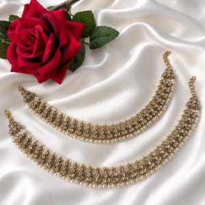 May include: A pair of gold-toned anklets featuring a repeating pattern of clear crystals and small white faux pearls. Each anklet has a delicate metal chain link design with a lobster claw clasp and an adjustable extender chain at the end.