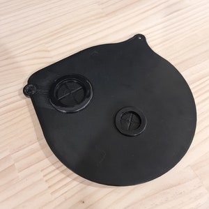 May include: A black, circular face mask with two circular filter vents. The mask has a rounded shape with a small loop at the top for attachment. The mask is made of a matte black material.