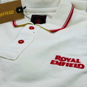 May include: White polo shirt with a red embroidered Royal Enfield logo. The collar has a red and white trim.