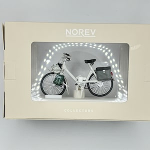 May include: A white model of a vintage bicycle with a green basket and a black saddle. The model is displayed in a clear plastic box with a white background. The box has the word "NOREV" printed on the top and "COLLECTORS" printed on the bottom.