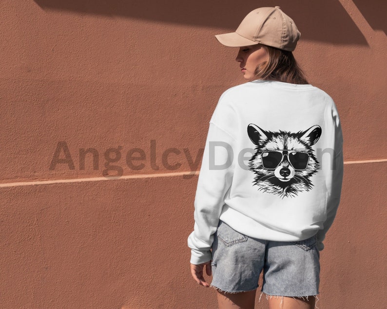 K&ouml;nnte beinhalten: Wei&szlig;es Sweatshirt mit einer schwarz-wei&szlig;en Illustration eines Waschb&auml;ren mit Sonnenbrille auf der R&uuml;ckseite. Der Waschb&auml;r schaut mit einem ernsten Ausdruck nach vorne.