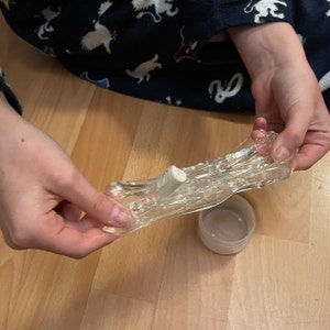 以下が含まれることがあります： 透明なスライムに虹色のグリッターと白い円柱が入っており、両手で伸ばして持たれています。スライムは透明な容器に入っています。