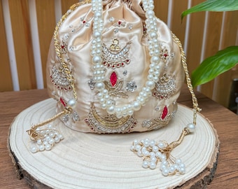 May include: A cream-colored drawstring bag with a pearl and gold beaded trim. The bag is decorated with intricate embroidery featuring red, gold, and silver accents.