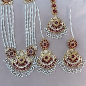 May include: A traditional Indian bridal jewellery set featuring a long pearl necklace with multiple strands, a matching pair of earrings, and a headpiece. The jewellery is adorned with intricate gold and red gemstone embellishments.