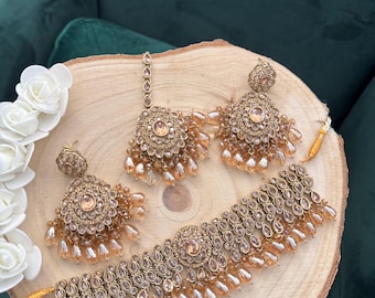 Pode incluir: Um conjunto de joias douradas com colar, brincos e um acessório de cabeça. O conjunto inclui um colar gargantilha, dois brincos redondos e um acessório de cabeça, todos adornados com pequenas pedras transparentes e contas penduradas em forma de gota. As joias são exibidas em uma superfície de madeira.