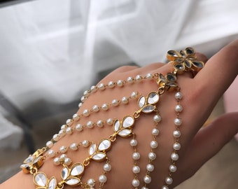 May include: A gold and white layered bracelet with pearl beads and floral accents. The bracelet is worn on a person's wrist and hand.