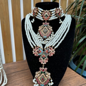 Könnte beinhalten: Ein mehrreihiges Perlenhalsband-Set mit einem großen, verzierten Anhänger mit bunten Edelsteinen. Der Anhänger hat die Form einer Blume mit filigranen Details. Die Halskette befindet sich auf einem schwarzen Samtständer.