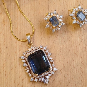 Peut inclure: Un collier en chaîne dorée avec un pendentif orné d'une pierre précieuse bleu foncé entourée de pierres de zircone cubique transparentes. Le pendentif est de forme rectangulaire avec une bordure décorative. Le collier est assorti à des boucles d'oreilles avec des pierres précieuses bleu foncé et des pierres de zircone cubique transparentes.