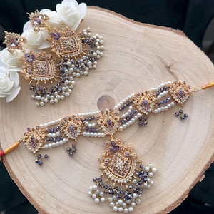 May include: A gold-toned jewelry set featuring a necklace and earrings, adorned with purple gemstones and white pearls. The set is displayed on a wooden surface, with white flowers in the background. The necklace has a central pendant.