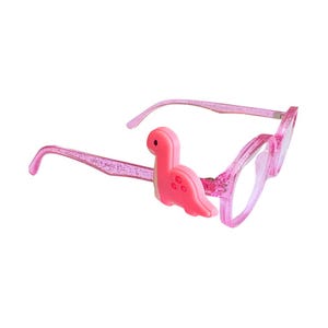 May include: Pink-framed eyeglasses with a glittery finish and a pink dinosaur accent on the left side. The glasses have clear lenses and a playful, kid-friendly design. The dinosaur is a bright pink color.