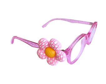 May include: Pink eyeglasses with a flower accent. The frames are a translucent pink with a glittery finish. A large, decorative flower with pink and white petals and a yellow center is attached to the frame.