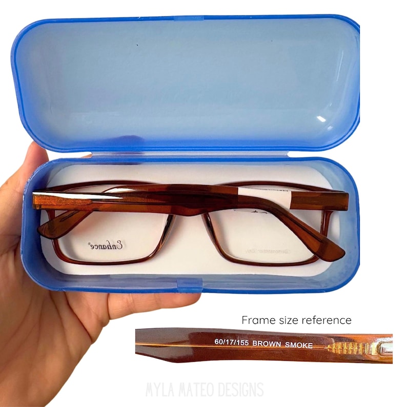 May include: Brown rectangular eyeglasses with the brand name Enhance printed on the lens, housed in a translucent blue plastic case. A detail view shows the frame size reference 60/17/155 and the color name Brown Smoke printed on the temple arm.