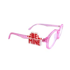 May include: Pink eyeglasses with clear lenses. The frames are a glittery pink and feature a white and red embellishment that reads "BE MINE". The glasses have a classic rectangular shape.