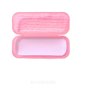 May include: A rectangular pink plastic case with a hinged lid. The interior of the lid features a textured pink lining, while the bottom section of the case has a smooth, light pink base. The text MYLA MATEO DESIGNS is printed at the bottom.
