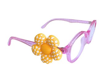 May include: Pink eyeglasses with a flower accent. The frames are a translucent pink with glitter. The flower is yellow and white with a checkered pattern. The lenses are clear.