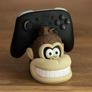 May include: A black and white Nintendo Switch Pro Controller rests on a cartoon monkey-shaped stand. The stand is tan and brown with a large, smiling mouth and white teeth. The background is a wooden surface.