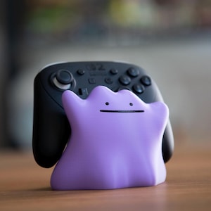 May include: A black video game controller with a purple Ditto-shaped stand. The controller has multiple buttons and joysticks. The Ditto stand is a light purple color with a simple face.