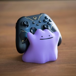 May include: A black Xbox controller with gold accents is paired with a purple Ditto figure. The controller has buttons and joysticks. The Ditto figure has a simple smiling face. The items are on a wooden surface.