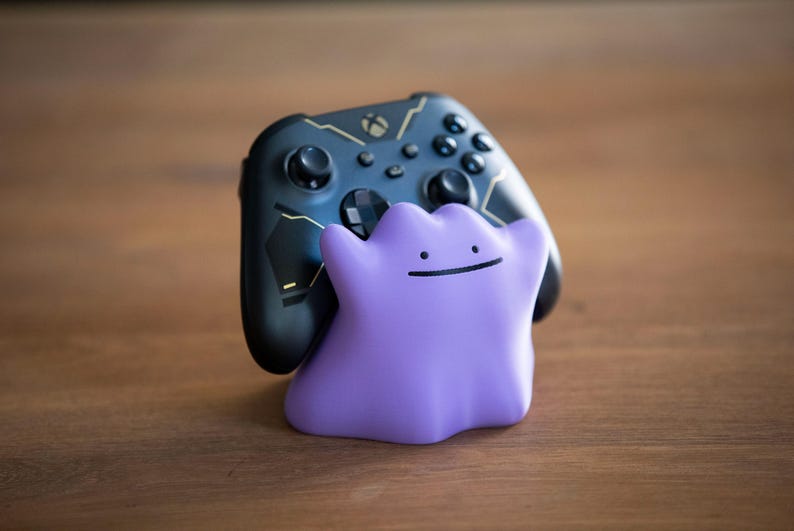 May include: A black Xbox controller with gold accents is paired with a purple Ditto figure. The controller has buttons and joysticks. The Ditto figure has a simple smiling face. The items are on a wooden surface.