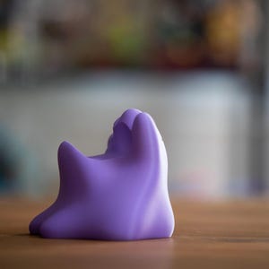 May include: A solid, lavender-colored, abstract sculpture with a rounded base and a textured, organic form. The object is positioned on a wooden surface, with a blurred background.