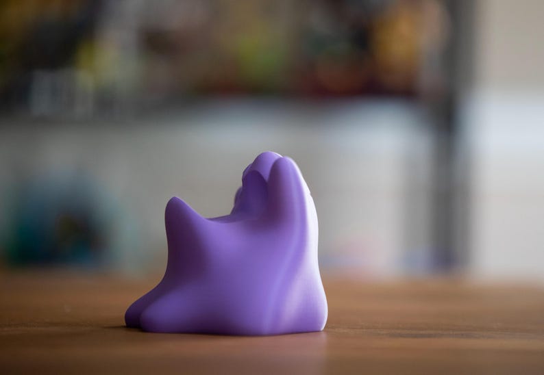 May include: A solid, lavender-colored, abstract sculpture with a rounded base and a textured, organic form. The object is positioned on a wooden surface, with a blurred background.