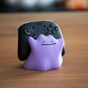 May include: A black Nintendo Switch Pro Controller with a purple Ditto-shaped stand. The controller has multiple buttons and a D-pad. The Ditto stand is a light purple color with a simple, smiling face.