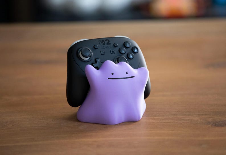 May include: A black Nintendo Switch Pro Controller with a purple Ditto-shaped stand. The controller has multiple buttons and a D-pad. The Ditto stand is a light purple color with a simple, smiling face.