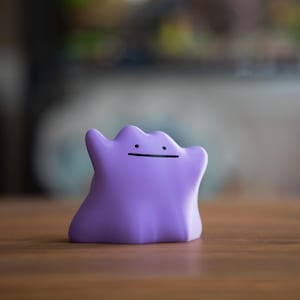May include: A small, purple Ditto figurine with a simple black line for a mouth. The figure is smooth and rounded, with two small arms raised. The background is blurred, with a wooden surface in the foreground.