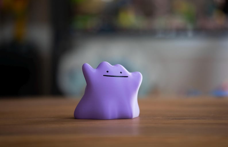 May include: A small, purple Ditto figurine with a simple black line for a mouth. The figure is smooth and rounded, with two small arms raised. The background is blurred, with a wooden surface in the foreground.