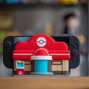 May include: A red and white 3D-printed accessory shaped like a building, designed to hold a black Nintendo Switch console. The building has a red roof with a white Poké Ball symbol and a blue door. The accessory is on a wooden surface.