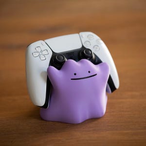 May include: A white and black PlayStation 5 controller rests on a purple Ditto-shaped stand. The stand has a simple smiling face. The items are on a wooden surface.
