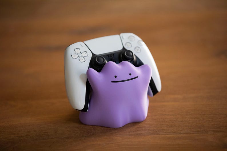 May include: A white and black PlayStation 5 controller rests on a purple Ditto-shaped stand. The stand has a simple smiling face. The items are on a wooden surface.