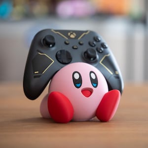 May include: A pink Kirby character figurine with a black and gold Xbox controller resting on its back. The controller is facing the camera.